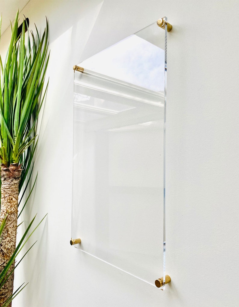 May include: A clear acrylic frame with gold metal mounting hardware. The frame is hanging on a white wall.