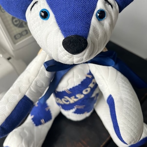 Könnte beinhalten: Ein weißer und blauer Patchwork-Teddybär mit blauen Augen und einer schwarzen Nase. Der Bär sitzt auf einer Holzoberfläche und hat den Namen "Jackson" auf seiner Brust gestickt.