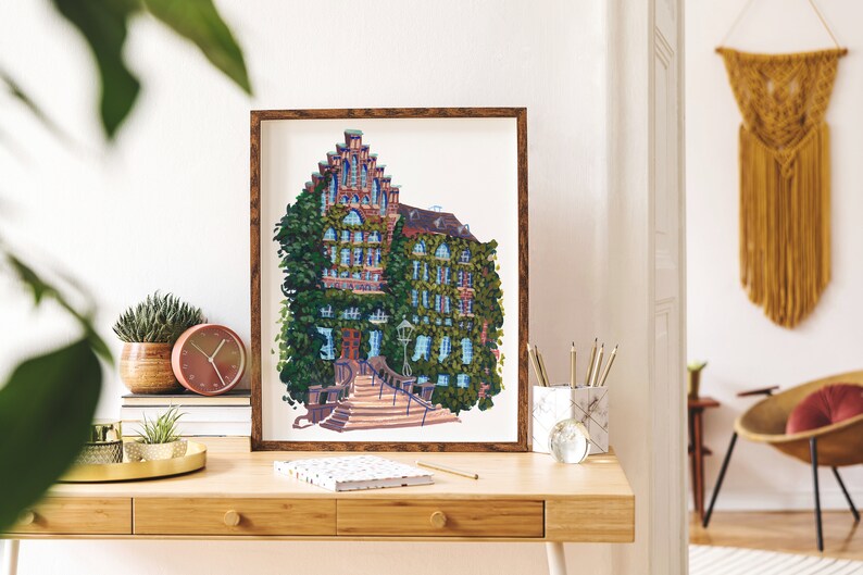 May include: A framed illustration of a brick building with a steep roof covered in green ivy. The building has a set of stairs leading up to the entrance. The illustration is in a brown frame and is hanging on a white wall.