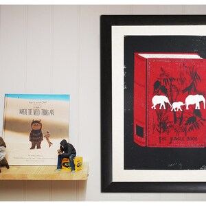May include: A framed black and white print of a red book with the title "The Jungle Book" and a white illustration of three elephants. The book is on a black background.