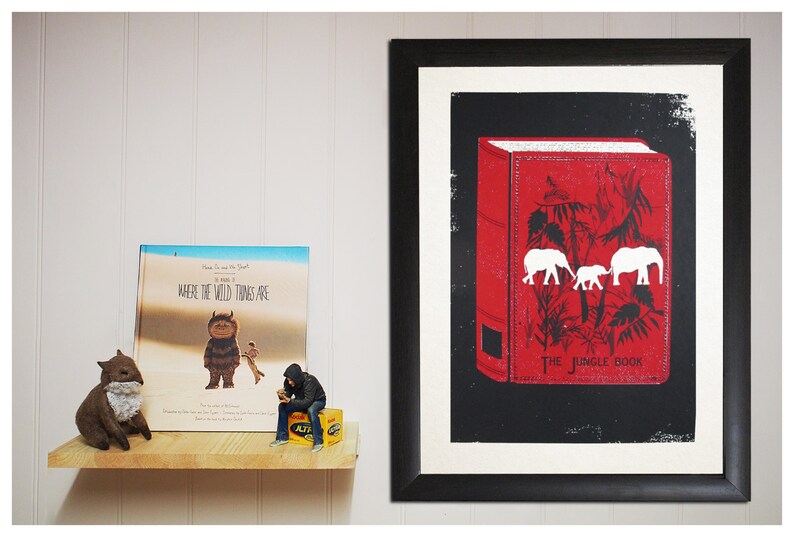 May include: A framed black and white print of a red book with the title "The Jungle Book" and a white illustration of three elephants. The book is on a black background.