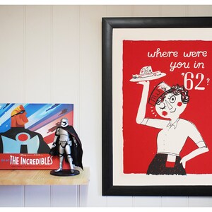 May include: A framed print with a red background and a black and white illustration of a woman holding a plate with a pie on it. The text on the print reads "where were you in '62?"