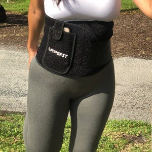 Könnte beinhalten: Schwarzer Taillentrainer mit einer Handytasche und dem aufgedruckten Text "LAPSFIT". Der Taillentrainer wird über einem weißen Tanktop und grauen Leggings getragen.