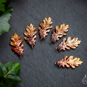 Könnte beinhalten: Sechs bronzefarbene Eichenblatt-Anhänger auf einer dunkelgrauen Oberfläche. Die Blätter haben detaillierte Adern und ein metallisches Finish, mit einem Farbverlauf von Gold zu Kupfer. Die Blätter sind verstreut, was ein natürliches, herbstliches Thema erzeugt.
