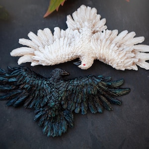 Könnte beinhalten: Zwei dekorative Vogelskulpturen, eine weiße und eine dunkelblaue. Beide haben ausgebreitete Flügel und sind vor einem dunklen Hintergrund mit Blättern platziert.