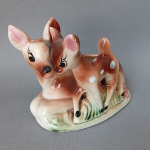 May include: A ceramic planter shaped like two deer, one larger than the other. The deer are brown with white spots and have pink noses. The planter is a light brown colour with green accents.