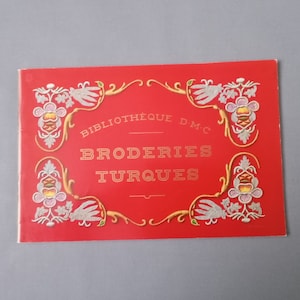 Op de afbeelding: Een rode boekomslag met gouden letters en decoratieve bloemen- en handachtige ontwerpen. De tekst luidt "BIBLIOTHEQUE D.M.C. BRODERIES TURQUES". De omslag heeft een vintage uitstraling.