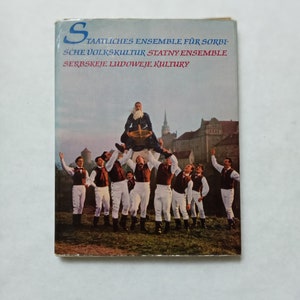 Può includere: Una copertina di libro vintage con un gruppo di persone in abiti tradizionali, che sollevano una persona con un cesto. La copertina presenta testo in più lingue. Sullo sfondo un castello e un campo verde.