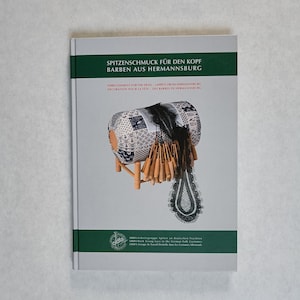 May include: A book titled "Spitzenschmuck für den Kopf Barben aus Hermannsburg" with a green banner. The cover features a detailed illustration of a lace head covering on a wooden stand. The book's title is in German, with English and French translations.