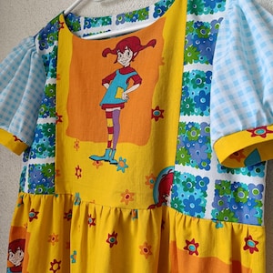以下が含まれることがあります： 漫画のキャラクタープリントが特徴的なカラフルなドレス。 ドレスは、花柄の黄色のボディスと、漫画のキャラクターが描かれた大きなオレンジ色のパネルを備えています。 袖は青と白のチェック柄で、ネックラインは黄色でトリミングされています。