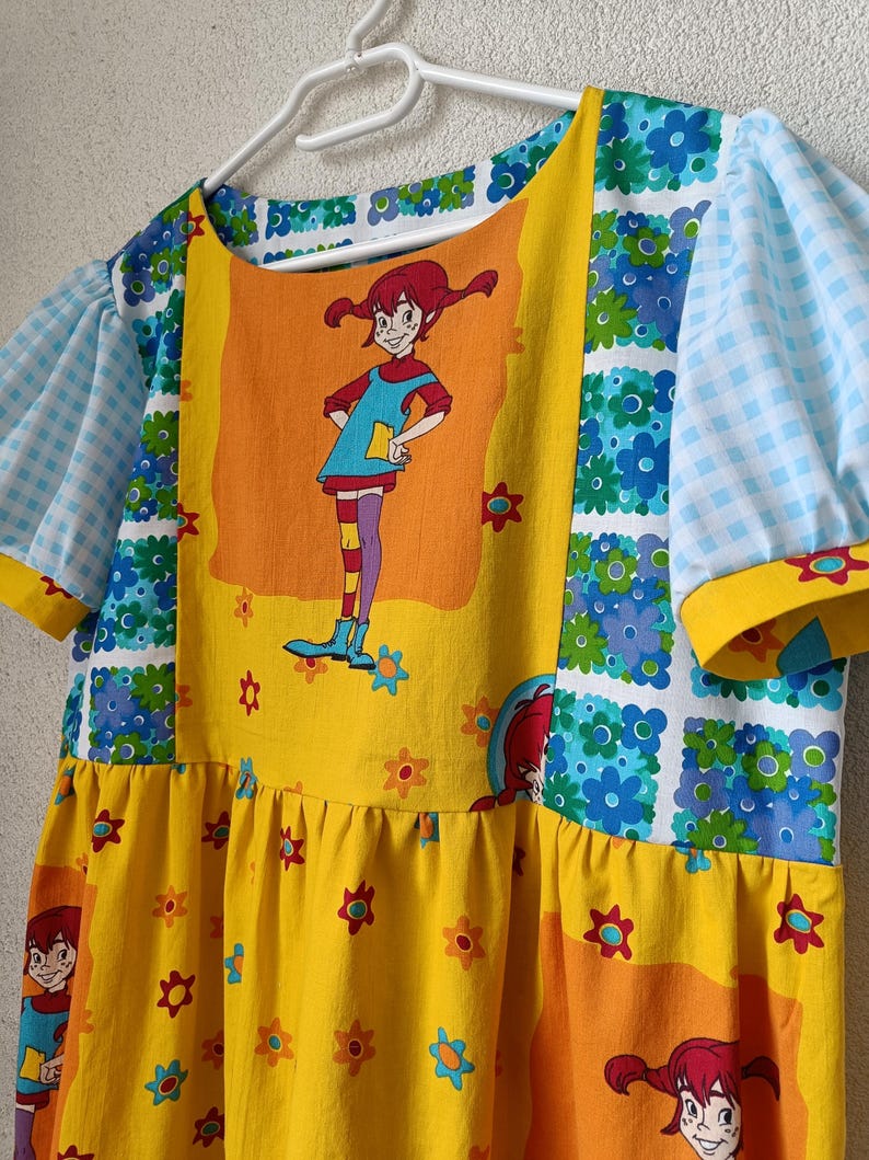 以下が含まれることがあります： 漫画のキャラクタープリントが特徴的なカラフルなドレス。 ドレスは、花柄の黄色のボディスと、漫画のキャラクターが描かれた大きなオレンジ色のパネルを備えています。 袖は青と白のチェック柄で、ネックラインは黄色でトリミングされています。