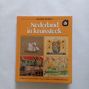May include: A yellow book titled "Nederland in kruissteek" by Henriette Beukers. The book cover features four cross-stitch designs: a ship, a coat of arms, a Dutch village, and a floral scene.
