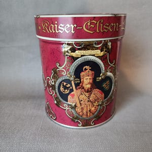 May include: A cylindrical red tin with gold lettering that reads "Kaiser-Elisen-". The tin features an illustration of a crowned figure holding a scepter, with ornate gold and black accents. The tin is likely a container for food items.