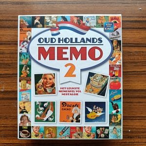 Puede incluir: Una caja cuadrada para el juego "Oud Hollands Memo 2" con un collage de anuncios vintage. La caja presenta la bandera holandesa y texto en holandés. La caja está sobre una superficie de madera.