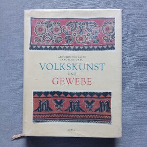 以下が含まれることがあります： 「Volkskunst und Gewebe」というタイトルの本。赤、青、黒の装飾的な花柄と幾何学模様が特徴です。表紙にはアントニス・ヴァツラフとヤロスラフ・オレルの名前が記載されています。
