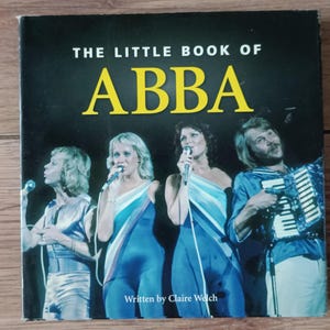Könnte beinhalten: Ein Buch mit dem Titel "The Little Book of ABBA" mit einem dunklen Einband und dem Bandnamen in großen gelben Buchstaben. Das Cover zeigt die vier ABBA-Mitglieder in blauen Outfits, die in Mikrofone singen. Der Name des Autors, Claire Welch, ist ebenfalls sichtbar.