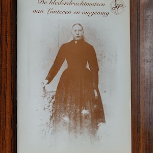 May include: A book cover featuring a sepia-toned photograph of a person in a dark dress. The title reads "De klederdrachtmutsen van Lunteren en omgeving" with a logo in the upper right corner. The background is a wooden surface.