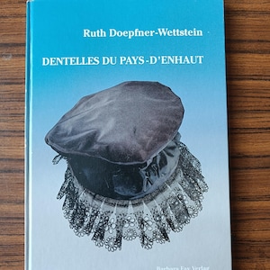 May include: A book titled "Dentelles du Pays-D'Enhaut" by Ruth Doepfner-Wettstein. The book cover features a black velvet hat with a black lace trim. The book is published by Barbara Fay Verlag.