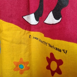 以下が含まれることがあります： 黄色と赤色の配色が施された生地のクローズアップ。 白い蹄を持つ黒い動物のデザインが特徴です。 生地には花の模様と「&copy; 1998 Iduna TFC Nelvana/SF」の文字も入っています。