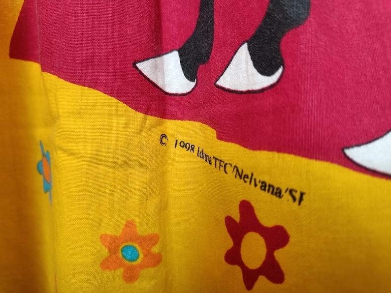 以下が含まれることがあります： 黄色と赤色の配色が施された生地のクローズアップ。 白い蹄を持つ黒い動物のデザインが特徴です。 生地には花の模様と「&copy; 1998 Iduna TFC Nelvana/SF」の文字も入っています。