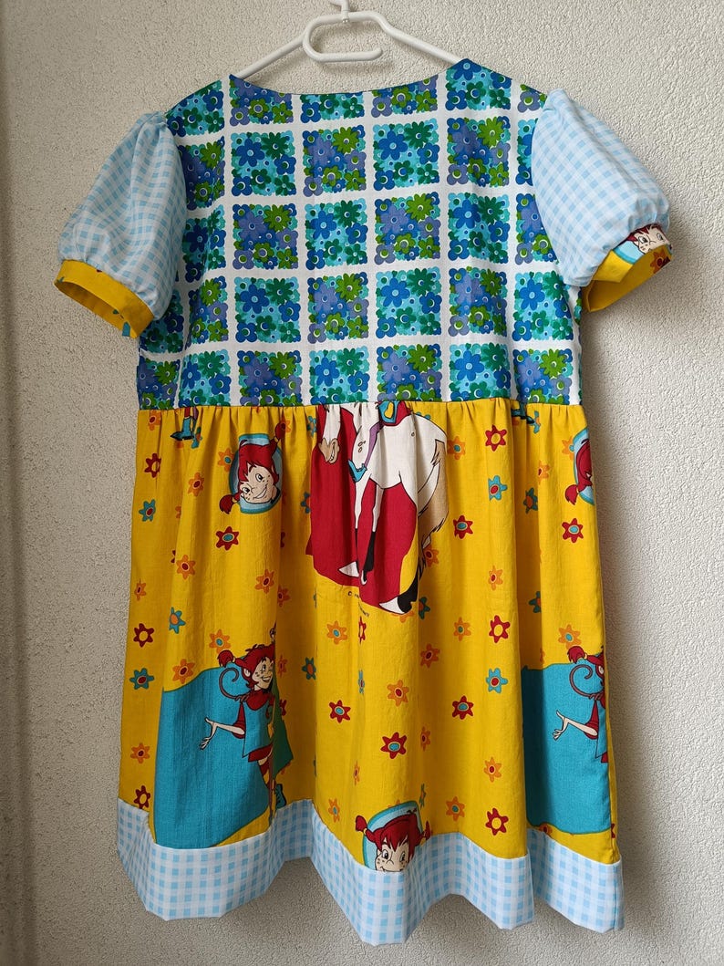以下が含まれることがあります： パッチワークデザインが特徴的なカラフルなドレス。上部は青と白の花柄で、袖は黄色のトリムが付いた水色のギンガムチェック。スカートは黄色で、漫画のキャラクターのイラストと青と白のギンガムチェックの裾が付いています。
