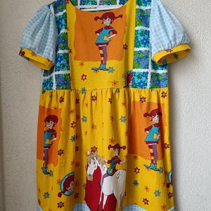 以下が含まれることがあります： 漫画のキャラクタープリントが施された、カラフルな半袖ワンピース。 ワンピースは、キャラクターのイラストと花のアクセントが付いた黄色のボディスです。 袖は水色のギンガムチェックで、ネックラインは花柄で縁取られています。 裾は青と白のギンガムチェックでトリミングされています。