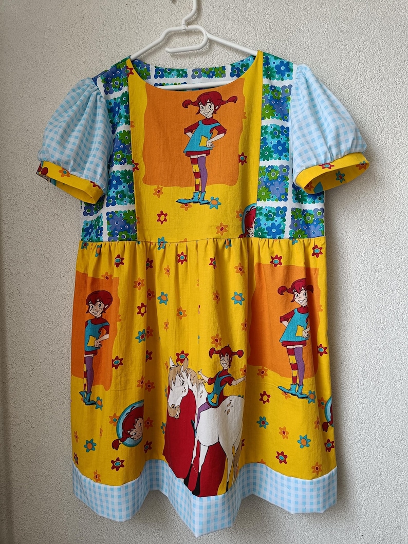 以下が含まれることがあります： 漫画のキャラクタープリントが施された、カラフルな半袖ワンピース。 ワンピースは、キャラクターのイラストと花のアクセントが付いた黄色のボディスです。 袖は水色のギンガムチェックで、ネックラインは花柄で縁取られています。 裾は青と白のギンガムチェックでトリミングされています。
