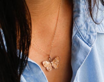 May include: A gold-colored butterfly pendant necklace. The butterfly pendant has detailed wing patterns and hangs from a delicate chain. The necklace is worn against a blue denim shirt.