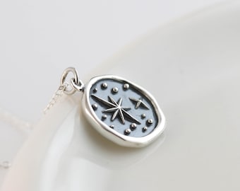 May include: A silver pendant necklace with an oval charm. The charm has a dark grey background with a starburst design and small star accents. The necklace is on a white surface.
