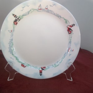 Puede incluir: Un plato de cena de cerámica blanca con un borde azul y verde que presenta un faro, veleros y gaviotas. El plato está en un soporte blanco.