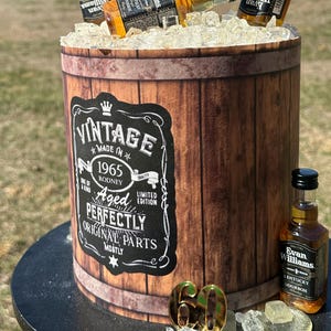 May include: A cake decorated to resemble a whiskey barrel, with the words "Aged to Perfection" on top. The cake is adorned with miniature Evan Williams bourbon bottles and ice. A "Vintage" label is on the side, with the number "60" in gold.