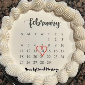 May include: A round cake decorated with white frosting and a calendar design for February. The calendar highlights the 14th with a red heart. The cake is also decorated with a border of white frosting swirls. The text "Your Optional Message" is at the bottom.
