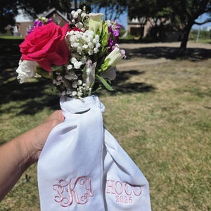 May include: A bouquet of flowers featuring a large, vibrant pink rose, white baby's breath, and purple and white blooms. The flowers are wrapped in a white fabric with pink embroidered initials and the text "HOCO 2025".