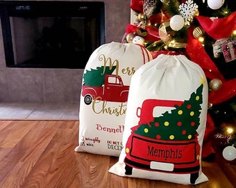 May include: Two off-white Christmas gift bags with red truck and Christmas tree designs. One bag has the text "Merry Christmas" and "Bennett" printed on it. The other bag has the word "Memphis" printed on it. A decorated Christmas tree is in the background.