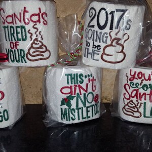May include: Six white toilet paper rolls with humorous Christmas-themed embroidery. The rolls are wrapped in clear plastic and tied with red ribbon. The text on the rolls includes "Santa's Tired of Your", "2017 is Going to be the", "Ho Ho Ho Gotta Go", "This Ain't No Mistletoe", "You're on Santa's List", and a poop emoji.
