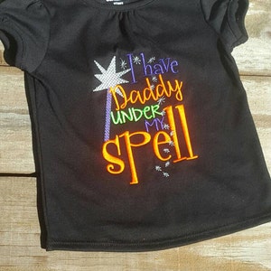 May include: Black short-sleeved shirt with a white, purple, and orange embroidered design that says "I have Daddy under my spell".