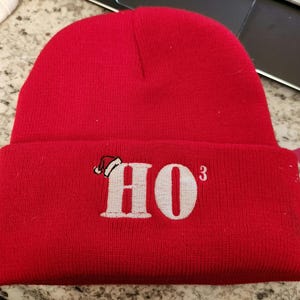Könnte beinhalten: Rote Strickmütze mit einem weißen gestickten Weihnachtsmann-Hut und dem Text "HO" mit einem Hochgestellt "3".