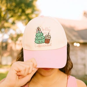 May include: A baseball cap with a light pink bill and a cream-colored crown. The cap features an embroidered design of a Christmas tree and a coffee cup with the words "Jolly and Caffeinated".