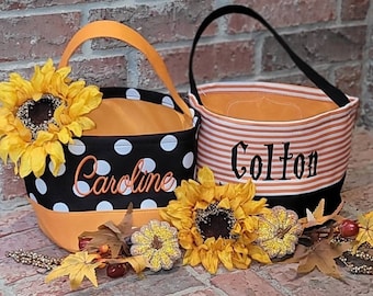 May include: Two orange and black Halloween trick-or-treat bags with the names "Caroline" and "Colton" embroidered on them. The bags are decorated with black and white polka dots and orange and white stripes. The bags are surrounded by fall foliage, including sunflowers.