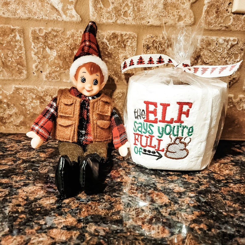 May include: A Christmas elf doll wearing a red and black plaid shirt and a brown vest sits next to a white toilet paper roll with the text "The Elf Says You're Full Of" embroidered on it.