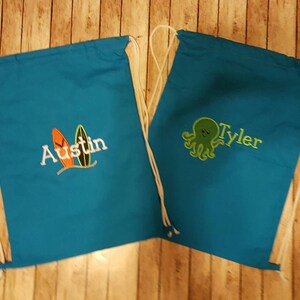 May include: Two blue drawstring bags with white drawstrings. One bag has a design of two surfboards and the name "Austin" embroidered on it. The other bag has a green octopus design and the name "Tyler" embroidered on it.