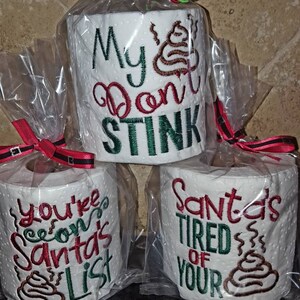May include: Three rolls of toilet paper wrapped in clear plastic with red and green ribbon. The toilet paper has embroidered designs with the text "My  Don't Stink", "You're  on Santa's List", and "Santa's Tired of Your" with a poop emoji on each roll.