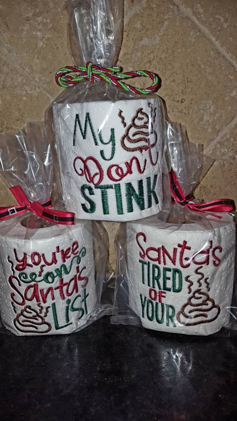 May include: Three rolls of toilet paper wrapped in clear plastic with red and green ribbon. The toilet paper has embroidered designs with the text "My  Don't Stink", "You're  on Santa's List", and "Santa's Tired of Your" with a poop emoji on each roll.