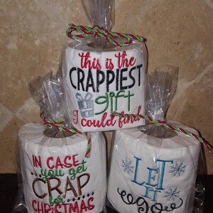 May include: Three rolls of toilet paper wrapped in clear plastic with red and green ribbon. The toilet paper rolls are decorated with embroidered text. The top roll says "This is the Crappiest gift I could find". The middle roll says "In case you get crap for Christmas". The bottom roll says "Let it go".