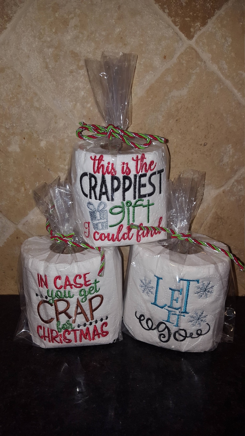 May include: Three rolls of toilet paper wrapped in clear plastic with red and green ribbon. The toilet paper rolls are decorated with embroidered text. The top roll says "This is the Crappiest gift I could find". The middle roll says "In case you get crap for Christmas". The bottom roll says "Let it go".
