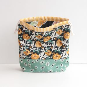 May include: A fabric drawstring bag with a floral pattern. The bag features a dark background with yellow flowers and green leaves. The bottom is a light green with white daisy-like flowers. The top edge has a yellow and white striped lining.