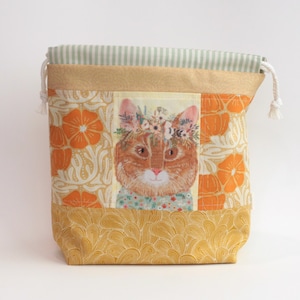 May include: A fabric drawstring pouch featuring a central panel with an orange cat wearing a floral crown. The bag has a light green striped top, a gold-toned band, and side panels with orange pumpkin motifs over a cream background.