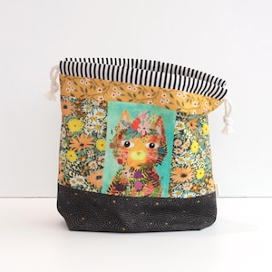 Puede incluir: Una bolsa con cordón con un colorido estampado floral y una ilustración de gato. La bolsa presenta una parte superior a rayas blancas y negras, una banda floral de color amarillo mostaza y una base negra estampada. Los cordones blancos completan el diseño.