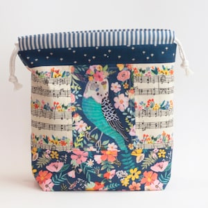 May include: A fabric drawstring pouch featuring a central panel with a budgie bird surrounded by flowers and musical notes. The bag has a dark blue top band with white stars, a striped blue and white trim, and a bottom section with a floral print.