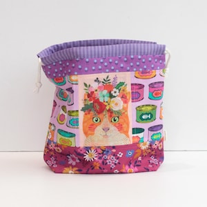 Puede incluir: Una bolsa con cordón con una ilustración de un gato con una corona floral. La bolsa presenta una combinación de colores púrpura y rosa con una cara de gato, latas de comida para gatos y estampados florales. La bolsa tiene un cordón blanco.